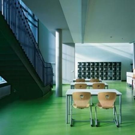 Helles, modernes Klassenzimmer mit grünem Bodenbelag, drei Holzstühlen, einer Treppe und Schließfächern